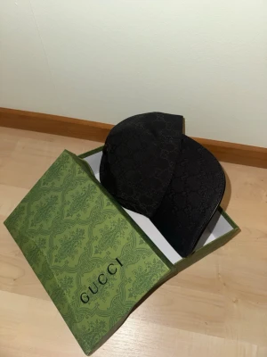 Svart keps från Gucci med monogram - Säljer en svart keps från Gucci med klassiskt GG-monogram i tyg. Kepsen har böjd skärm och justerbart band bak. Kommer i original grön Gucci-box. Perfekt accessoar för dig som gillar exklusiv streetwear.