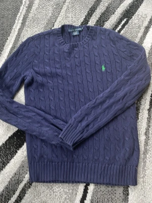 Manrinblå  kabelstickad tröja Polo Ralph Lauren - Säljer en mörkblå kabelstickad tröja från Polo Ralph Lauren med rund halsringning och grönt broderat logotyp på bröstet. Tröjan har långa ärmar och ribbade muddar vid ärmslut och nederkant. Perfekt för en stilren och klassisk look.