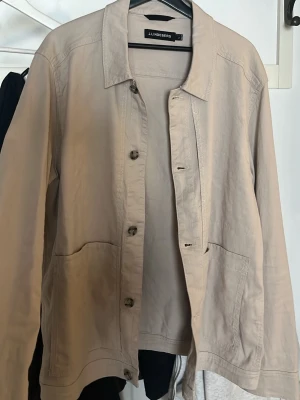 Beige overshirt från J.Lindeberg - Stilren beige overshirt från J.Lindeberg med klassisk krage, knäppning framtill och två stora fickor. Tillverkad i mjukt bomullsmaterial och har långa ärmar med knapp vid ärmslut. Perfekt för lager på lager och enkel att matcha.