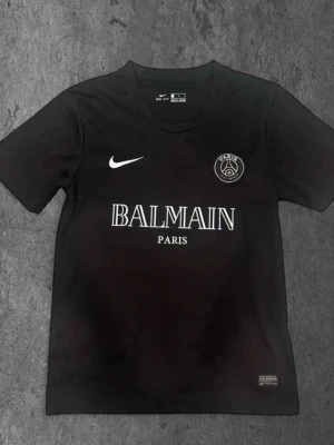 Svart PSG x Balmain t-shirt Nike - Säljer en svart t-shirt från Nike med Paris Saint-Germain x Balmain Paris tryck på bröstet. Tröjan har rund hals, korta ärmar och är tillverkad i Dri-FIT material som håller dig torr. Perfekt för dig som gillar exklusiva samarbeten och sportmode.