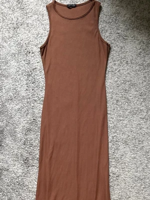 Ribbad brun långklänning - Säljer en ribbad brun långklänning med ärmlös design och rund halsringning. Klänningen har en slim passform och är tillverkad i ett mjukt, stretchigt material som sitter snyggt på kroppen. Perfekt för dig som gillar enkel och stilren look.
