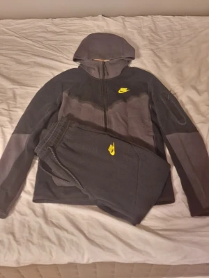 Mörkgrått Nike set med huva - Säljer ett mörkgrått set från Nike med både hoodie och matchande byxor. Setet har dragkedja framtill, huva och gula Nike-loggor på både överdel och underdel. Tillverkat i mjukt fleecematerial som känns riktigt skönt mot huden.