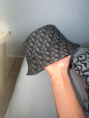 Dior bucket hat - Snygg mörkgrå bucket hat med all-over monogrammönster. Hatten har en klassisk form med mjuk, nedåtlutande brätte och är tillverkad i ett tyg med struktur. Perfekt för dig som vill ha en trendig accessoar som sticker ut.