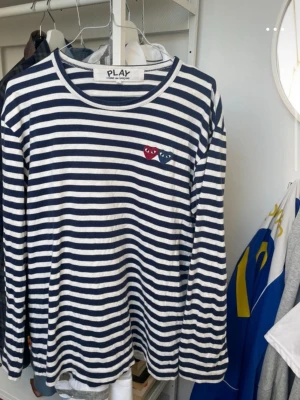 Randig långärmad tröja Comme des Garçons - Säljer en långärmad tröja från Comme des Garçons PLAY med marinblå och vita ränder. Tröjan har två hjärtapplikationer på bröstet, ett rött och ett blått. Tillverkad i mjuk bomull och har en avslappnad passform med rund hals.