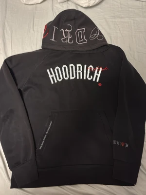 Hoodrich hoodie  - Svart hoodie från Hoodrich med stor vit logga på bröstet och broderade detaljer i rött och vitt. Huva med tryck och ficka framtill. Textdetaljer även på ärmen och ryggen. Tillverkad i mjukt bomullsmaterial för en skön och avslappnad look.