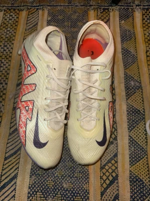 Nike Mercurial Zoom elite  fotbollsskor - Säljer ett par Nike Mercurial Zoom fotbollsskor i ljusgul färg med lila och röda detaljer. Skorna har snörning, textilöverdel och en sula med lila och orange mönster samt Zoom Air-teknologi för extra studs. Perfekta för dig som vill ha grymt grepp och lätt känsla på planen.