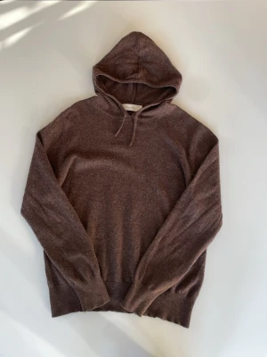 Brun kashmirhoodie från Sayless - Mysig brun hoodie från Soft Goat i mjuk och exklusiv kashmirblandning. Klassisk modell med huva och dragsko, samt ribbade muddar vid ärmslut och nederkant. Perfekt för dig som gillar stilrena och bekväma plagg.