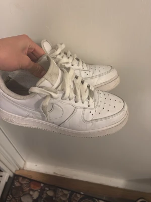 Vita Nike Air Force 1 sneakers - Klassiska vita Nike Air Force 1 sneakers med lågt skaft. Skorna har perforerad tå, vita skosnören och den ikoniska swoosh-loggan på sidan. Tillverkade i läder med gummisula för bra grepp. Perfekta för en clean och stilren look.