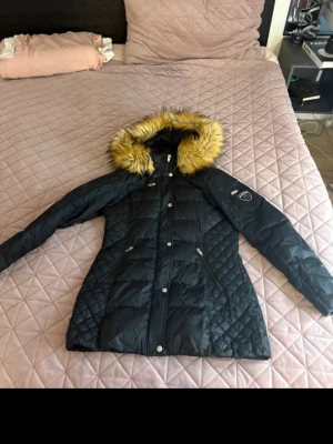 Svart dunjacka med päls från Rockandblue - Svart dunjacka från Rockandblue med quiltade detaljer och snygg passform. Jackan har en huva med fluffig, ljusbrun päls och flera fickor med dragkedja. Perfekt för kalla vinterdagar och riktigt stilren look.