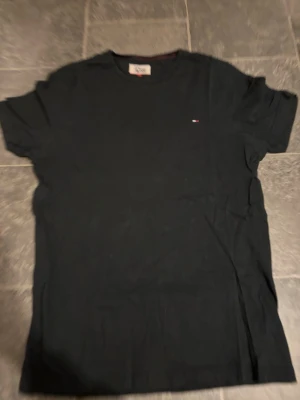 Svart t-shirt från Tommy Hilfiger - Klassisk svart t-shirt från Tommy Hilfiger Denim med liten logga på bröstet. T-shirten har rund halsringning och korta ärmar. Tillverkad i mjuk bomull som känns skön mot huden. Perfekt basplagg till garderoben.