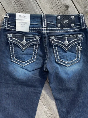 Miss Me bootcut jeans - Snygga mörkblå bootcut jeans från Miss Me med kontrasterande vita sömmar och detaljerade bakfickor med broderi och dekorativa knappar. Jeansen har låg midja och slitningar för en cool look. Tillverkade i denim med stretch för skön passform. Innersöm ben 86cm, rak över midja 36cm.