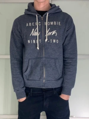 Blå zip-hoodie Abercrombie New York - Mysig blåmelerad zip-hoodie från Abercrombie med tryck 'New York Ninety-Two' på bröstet. Tröjan har huva med vita dragsnören, två fickor fram och ribbade muddar. Perfekt för en avslappnad och cool stil. Tröjan ska vara storlek L men sitter som S. Jag har fotat mått på bilderna. Jag är 183 och den sitter lite kort