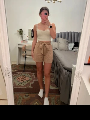 Beige highwaist shorts med knytband - Säljer ett par beige highwaist shorts med knytband i midjan. De har en lös passform och är tillverkade i ett mjukt bomullsliknande material. Perfekta för varma dagar och har en stilren look med fickor på sidorna.