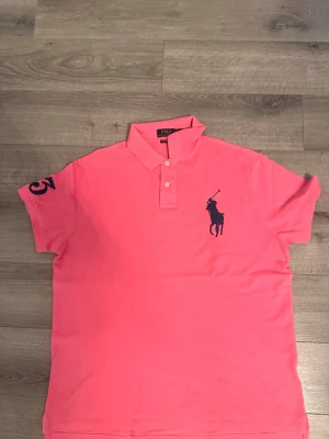Rosa Polo Ralph Lauren t-shirt - Säljer en rosa t-shirt från Polo Ralph Lauren med mörkblå logga på bröstet och siffran 3 på ärmen. Modellen är custom slim fit och har klassisk pikékrage med knappar. Perfekt för dig som gillar stilrena plagg med sportig touch.