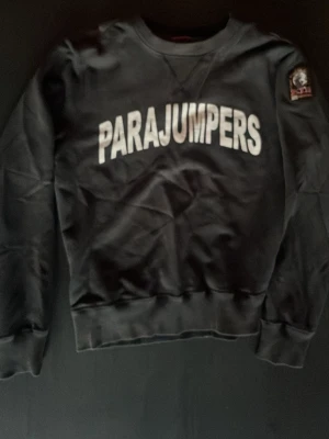 Svart sweatshirt från Parajumpers - Cool svart sweatshirt från Parajumpers med stor vit logga över bröstet och patch på ärmen. Tillverkad i mjuk fleece och har klassisk rund hals samt ribbade muddar vid ärmslut och nederkant. Perfekt för en avslappnad och trendig look.
