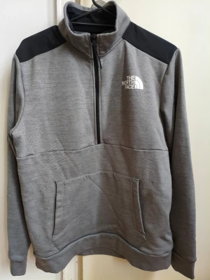 Grå half-zip tröja The North Face - Grå tröja från The North Face med svart axelparti och vit logga på bröstet. Modellen har half-zip dragkedja, hög krage och en stor magficka. Tillverkad i mjukt material och har långa ärmar. Perfekt för en avslappnad och sportig look.