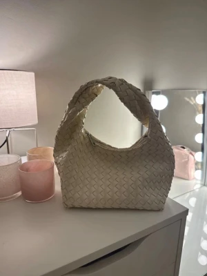 Vit flätad handväska med axelrem - Snygg vit handväska i flätad design med mjuk struktur och bred axelrem. Väskan har en modern, rundad form och är tillverkad i konstläder. Perfekt för dig som gillar trendiga accessoarer och vill ha en stilren look.