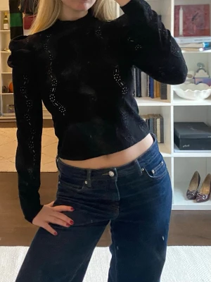 Svart långärmad topp med mönster och lite vidare axlar - Svart långärmad topp med höghals och vågformat hålmönster. Lätt puffade axlar och volangstuk. 