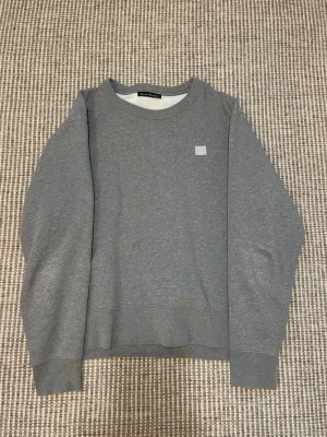 Grå sweatshirt från Acne Studios - Säljer en stilren grå sweatshirt från Acne Studios, modell Fairview Face. Tröjan har rund halsringning, lång ärm och en liten broderad Face-logga på bröstet. Tillverkad i 100% bomull för en mjuk och bekväm känsla.