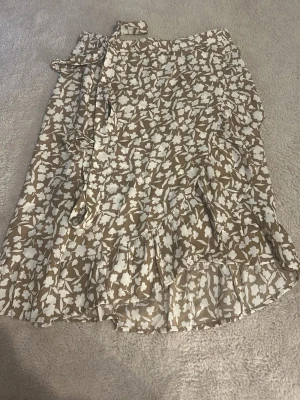 Beige blommig omlottkjol med volang - Säljer en lite mörkare beige omlottkjol med vitt blommönster och volangkant. Kjolen har knytband i midjan och är perfekt för dig som gillar en feminin och somrig stil. Lätt och luftig, passar till många olika outfits.
