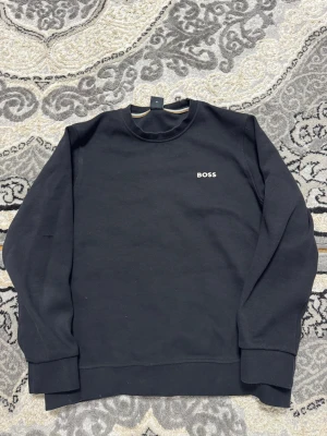 Svart stickad tröja från BOSS - Snygg svart stickad tröja från BOSS med rund halsringning och diskret BOSS-logga broderad på bröstet. Tröjan har långa ärmar och breda muddar vid ärmslut och nederkant. Perfekt för en clean och stilren look.