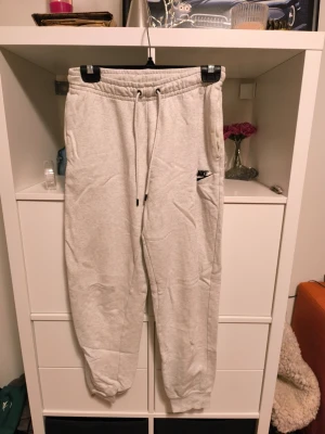 Gråa mjukisbyxor från Nike - Säljer ett par ljusgrå mjukisbyxor från Nike med broderad svart logga på benet. Byxorna har dragsko i midjan och muddar vid bensluten. Perfekta för chill dagar eller när du vill ha det extra bekvämt.