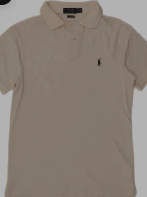 Beige pikétröja från Polo Ralph Lauren - Klassisk beige pikétröja från Polo Ralph Lauren med korta ärmar och krage. Tröjan har två knappar framtill och den ikoniska gröna logotypen broderad på bröstet. Perfekt till jeans eller shorts för en clean och stilren look.