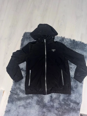 Svart vindjacka från Prada med huva - Snygg svart vindjacka från Prada med huva och dragkedja framtill. Jackan har två fickor med dragkedja och diskret Prada-logga på bröstet. Tillverkad i ett glansigt, lätt material som står emot vind. Perfekt för dig som vill ha en stilren och exklusiv look.