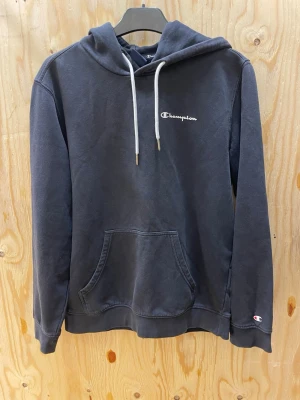 Mörkgrå hoodie från Champion - Mörkgrå hoodie från Champion med vit broderad logga på bröstet och liten logga på ärmen. Klassisk känguruficka framtill och vita dragsnören i huvan. Tillverkad i mjuk bomullsblandning för skön känsla och avslappnad stil.