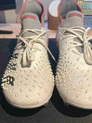 Adidas Predator fotbollsskor vita Elite - Säljer ett par Adidas Predator fotbollsskor i vitt med grå och röda detaljer. Skorna har en unik struktur med gummitaggar på ovandelen för extra grepp på bollen och en sprucken effekt i materialet. Klassisk snörning och låg profil, perfekt för dig som vill sticka ut på planen.