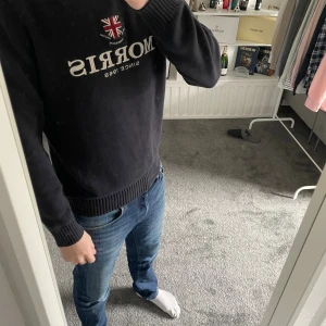 Morris tröja mörk blå - Snygg svart stickad tröja från Morris med rund halsringning och broderad logga samt brittisk flagga på bröstet. Tröjan har ribbade muddar vid ärmslut och nederkant. Perfekt för dig som gillar klassisk och stilren design.