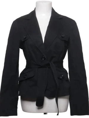 Svart trenchcoat med knytskärp - Stilren svart trenchcoat med klassisk krage, knytskärp i midjan och dekorativa knappar på bröstet och fickorna. Jackan har långa ärmar och två stora fickor framtill. Perfekt för dig som gillar en tidlös look.