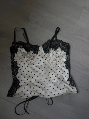 Prickig korsett med spetsdetaljer - Säljer en vit korsett med svarta prickar och tunna axelband. Korsetten har svarta spetsdetaljer längs bysten och sidorna samt bygel för extra stöd. Materialet är glansigt och ger en lyxig känsla. Perfekt för dig som vill ha något unikt och snyggt! Justerbar i ryggen