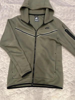 Nike tech - Snygg olivgrön hoodie från Nike med dragkedja framtill och svarta detaljer. Tröjan har huva, två bröstpaneler med reflexränder och en ficka med dragkedja på ärmen. Perfekt för en sportig och avslappnad stil.