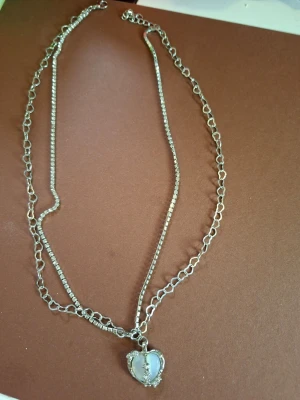 Silverfärgat halsband med hjärthänge - Dubbelkedjat halsband i silverfärg med ett hjärtformat hänge dekorerat med små pärlor. Ena kedjan har större länkar och den andra är tunnare och mer diskret. Perfekt accessoar för att lyfta din outfit.