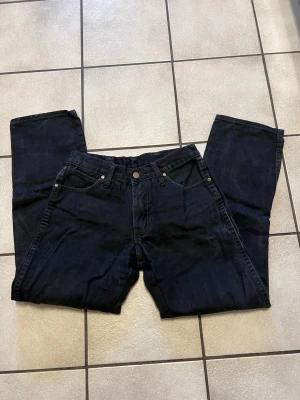 Svarta jeans från Wrangler storlek S, midja 68 cm - Säljer ett par svarta jeans från Wrangler med klassisk femficksdesign och raka ben. Jeansen har detaljer i metall och ett brunt lädermärke bak på ena fickan. Materialet är kraftig denim som ger en tidlös look.