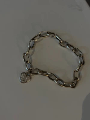 Länkarmband med hjärthänge - Snyggt armband i silver med grova länkar och ett gnistrande hjärthänge täckt av små stenar. Perfekt accessoar för att lyfta din outfit med lite extra bling. Passar dig som gillar trendiga och stilrena smycken. Märke: Thomas sabo