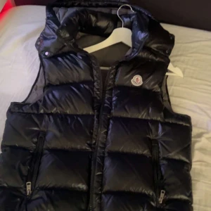 Svart dunväst från Moncler - Säljer en svart, glansig dunväst från Moncler med hög krage och dragkedja framtill. Västen har två fickor med dragkedja och Moncler-logga på bröstet. Perfekt för lager-på-lager under kyliga dagar.