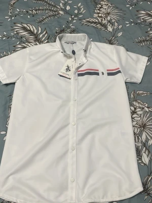 Vit skjorta från U.S. Polo Assn. - Vit skjorta med kort ärm från U.S. Polo Assn. Skjortan har knappar framtill, smal passform och dekorativa ränder i rött och blått över bröstet. Kragen har ett diskret svartvitt mönster på insidan och en liten logga på bröstet.