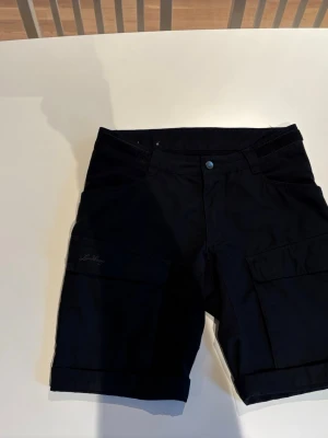 Svarta shorts från Lundhags - Säljer ett par svarta shorts från Lundhags med klassisk design och praktiska fickor på sidorna. De har en knapp framtill och är perfekta för dig som gillar outdoor och friluftsliv. Materialet känns slitstarkt och bekvämt.