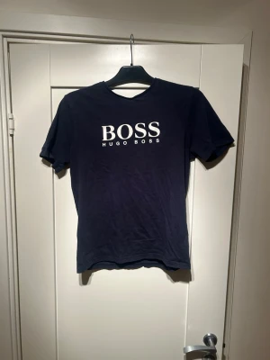 Mörkblå Hugo Boss t-shirt - Snygg mörkblå t-shirt från Hugo Boss med vit logga tryckt på bröstet. Klassisk passform med rund hals och korta ärmar. Tillverkad i mjuk bomull som känns skön mot huden. Perfekt för en clean och stilren look.
