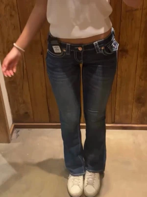 Blå bootcut jeans från True Religion - Säljer ett par mörkblå bootcut jeans från True Religion med tydliga vita kontrastsömmar och klassiska fickdetaljer. Jeansen har låg midja och är tillverkade i denim med stretch för skön passform. Perfekta för en trendig och avslappnad look.