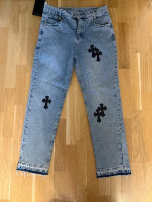 Ljusblå jeans med svarta korspatchar - Unika ljusblå jeans från Chrome Hearts med flera svarta korspatchar på fram- och baksidan. Jeansen har rak passform, klassisk femficksdesign och råa, fransiga benslut för en edgy look. Tillverkade i denim med cool streetwear-vibe.