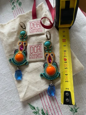 Färgglada örhängen från Dori Csengeri köpta på Svensk Tenn - Unika statementörhängen från Dori Csengeri med broderade detaljer i turkos, orange, lila och blått. Stora orange pärlor i mitten, lila glittrande stenar och blå droppformad pärla längst ner. Handgjorda och riktigt iögonfallande accessoarer.