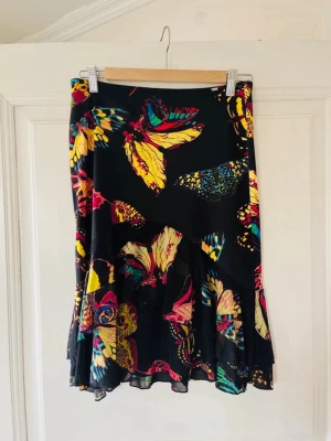 Y2k butterfly midiskirt with ruffled hem Carrie Bradshaw vibe - Unik svart kjol med färgglatt fjärilsmönster i gult, rött, blått och grönt. Kjolen har volangdetaljer och en lätt utsvängd form som ger en snygg siluett. Perfekt för dig som vill sticka ut med en cool och lekfull stil.