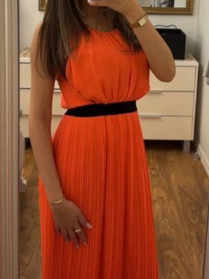 Plisserad orange maxiklänning - Snygg orange maxiklänning med plisserad kjol och svart midjeband. Ärmlös modell med rund halsringning och lös passform upptill. Perfekt för dig som vill sticka ut med färg.