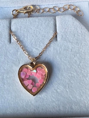 Halsband med hjärthänge och rosa detaljer - Sött halsband med ett hjärtformat hänge i guldfärgad metall. Hänget har en transparent mitt fylld med små rosa hjärtformade dekorationer som ger en lekfull och trendig look. Perfekt accessoar för dig som gillar gulliga och unika smycken.