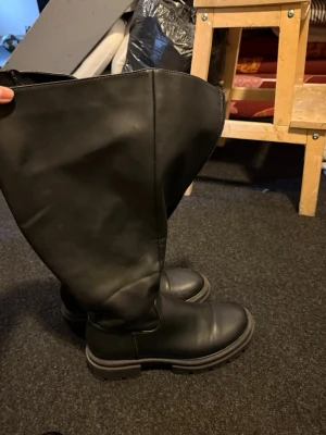 Svarta höga boots med grov sula - Säljer ett par svarta höga boots med dragkedja på sidan och rejäl grov sula. Stövlarna är i slätt konstläder och har en chunky look som passar till många olika stilar. Perfekta för dig som gillar en tuffare vibe på dina skor.