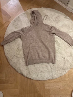 Beige kaschmir hoodie med huva - Mysig beige kaschmir hoodie med huva och dragsko. Tröjan har långa ärmar och ribbade muddar vid ärmslut och nederkant. Perfekt för chill dagar eller när du vill ha något bekvämt och stilrent. Materialet känns mjukt och skönt mot huden.