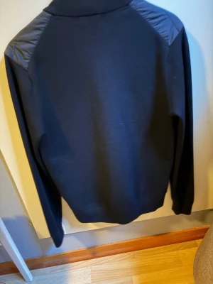 Svart cardiganjacka från J.Lindeberg - Snygg svart cardiganjacka från J.Lindeberg med quiltad framsida och stickade ärmar. Jackan har dragkedja framtill, ståkrage och två fickor. Perfekt nu till våren o sommaren, STRL S, Köpt i butik för 3000kr men använd 3 gånger bara 